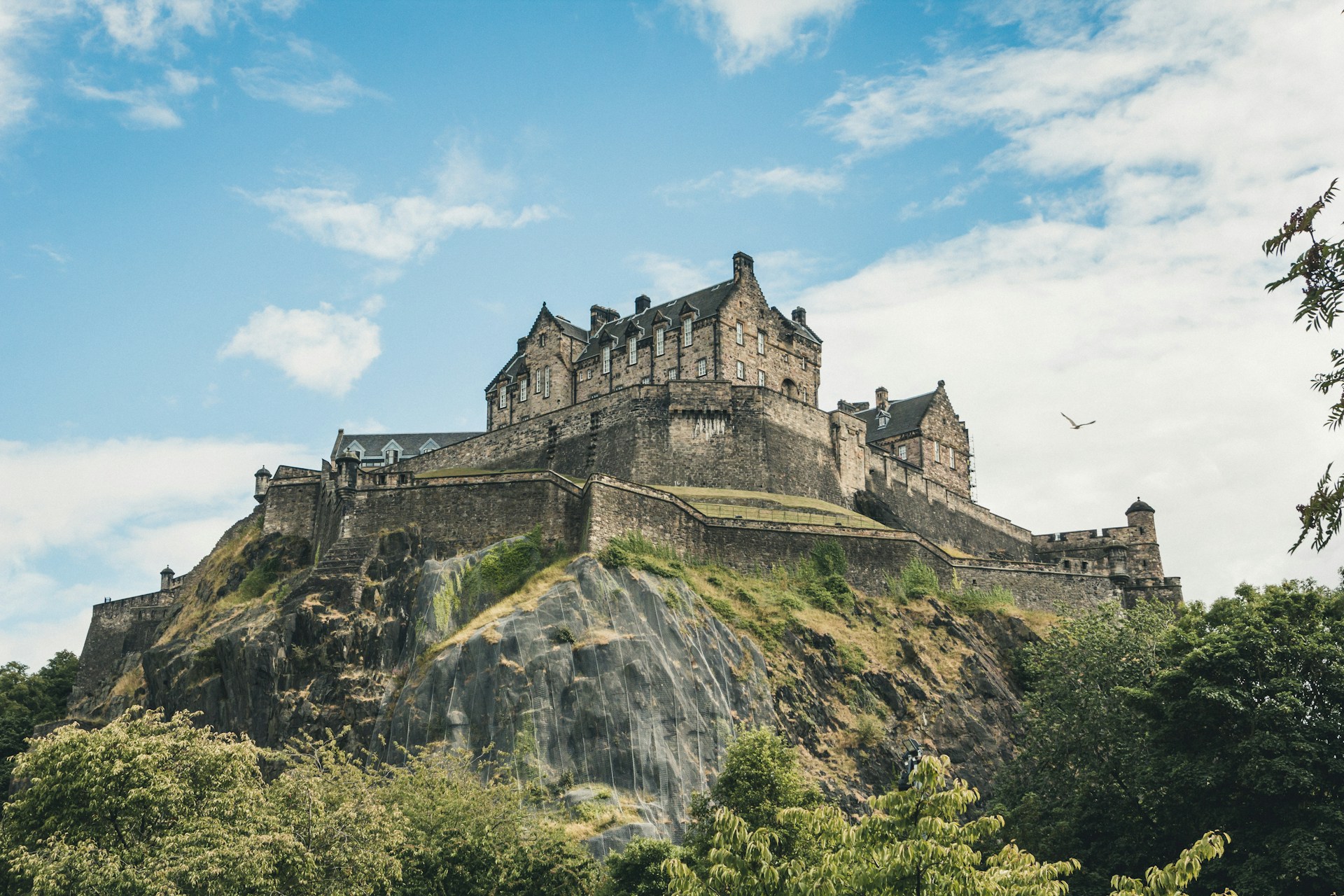 The stately Edinburgh Castle sitting on top of Castle Rock, surrounded by blue skies above and lush greenery below.