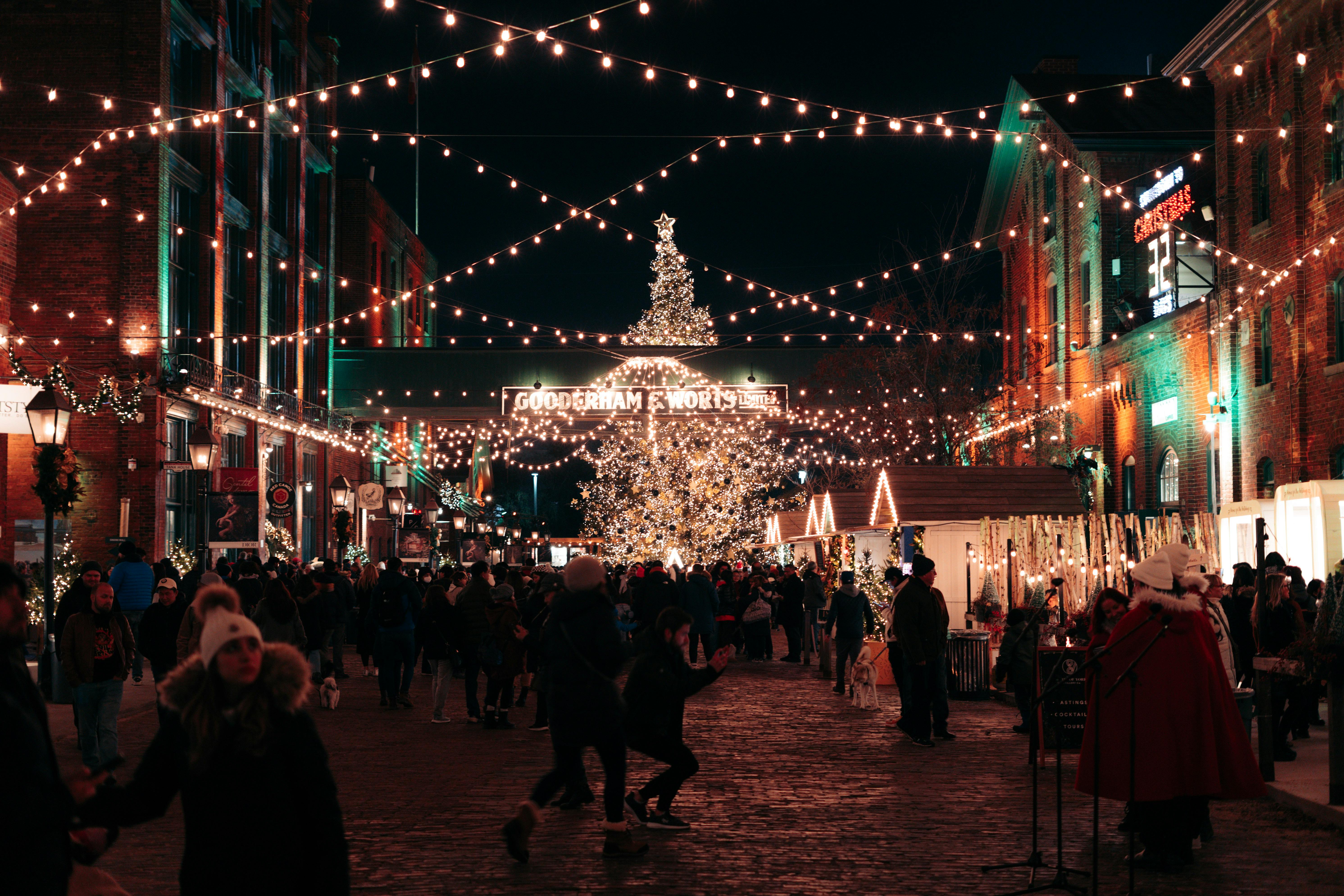 A lively winter market scene at night, with people strolling among festively lit stalls against a backdrop of glowing lights and seasonal decorations