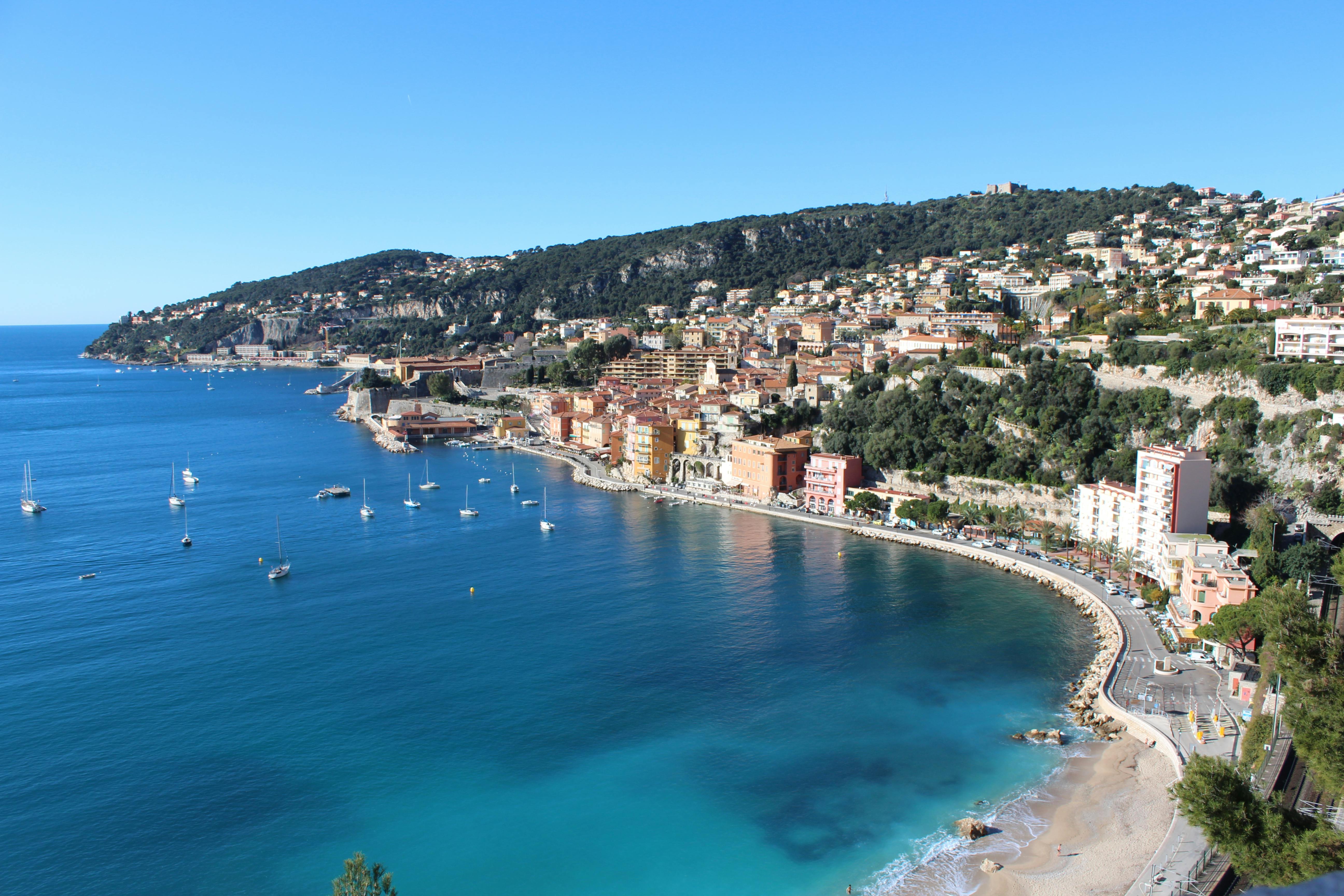 Elegant pastel-hued buildings line the waterfront, reflecting in the calm blue waters of the French Riviera under clear skies