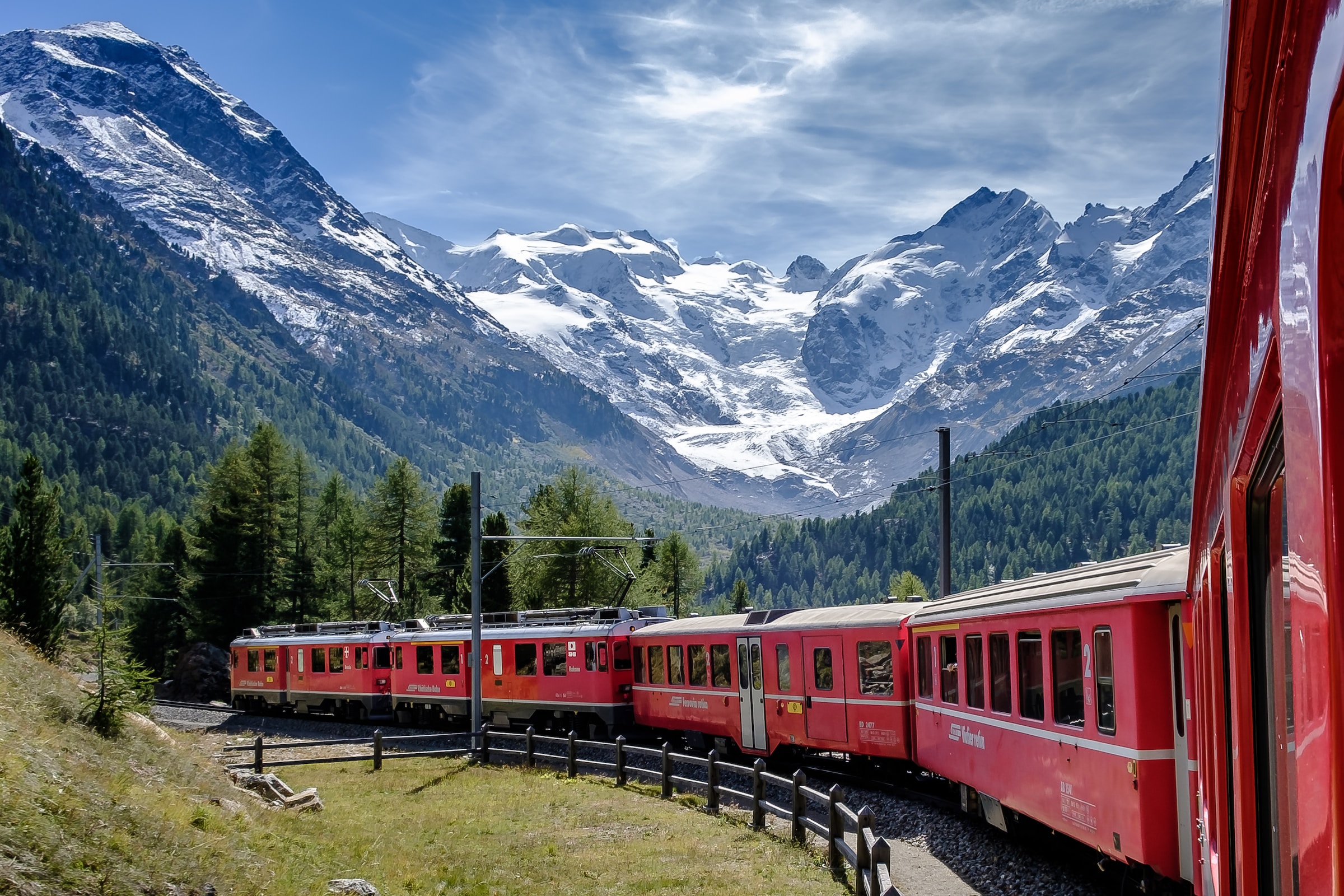 A red train passes through a range of mountains and trees
