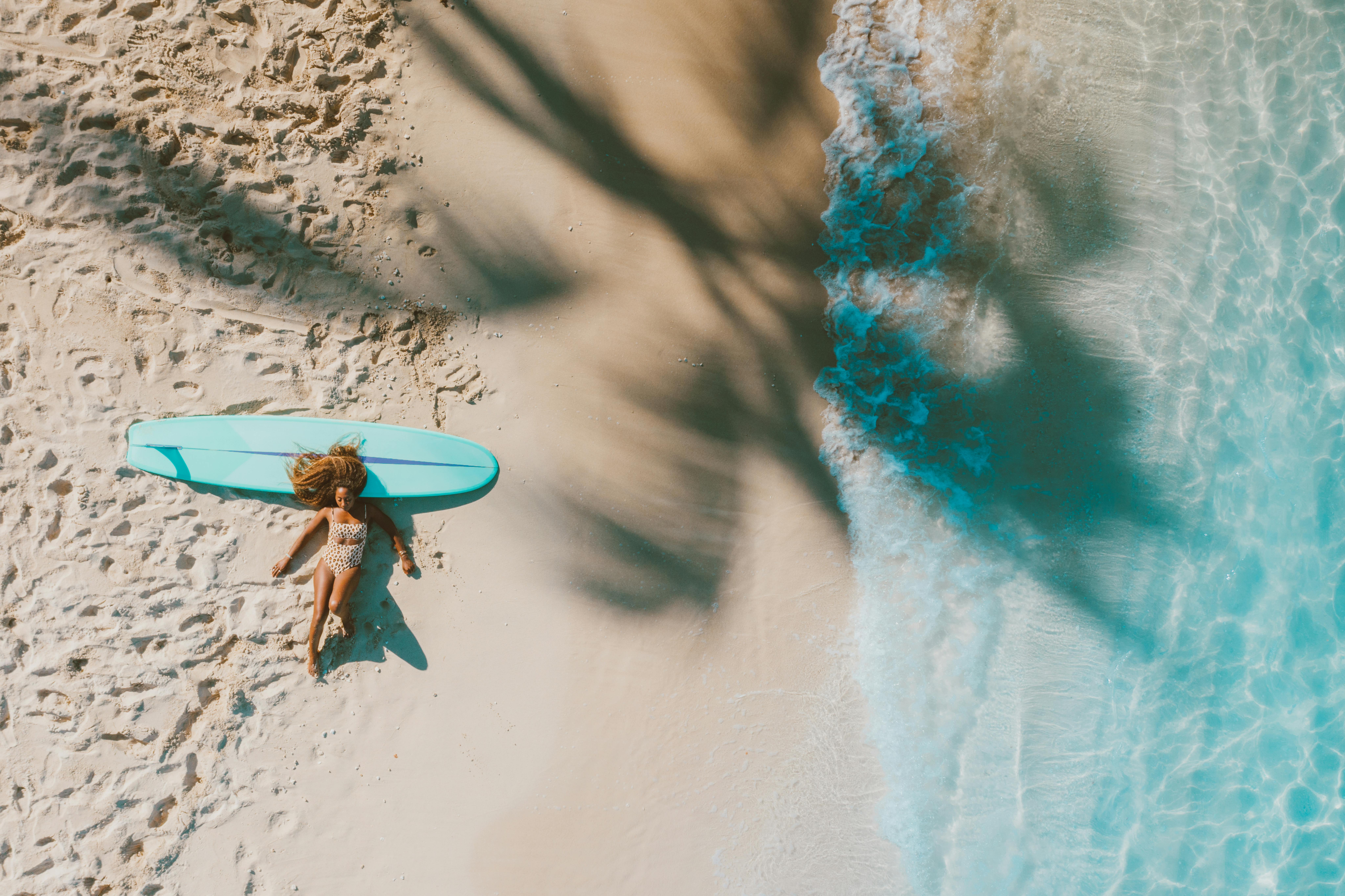 A top-view shot of a woman relaxing and sunbathing on her surfboard on a sandy beach.