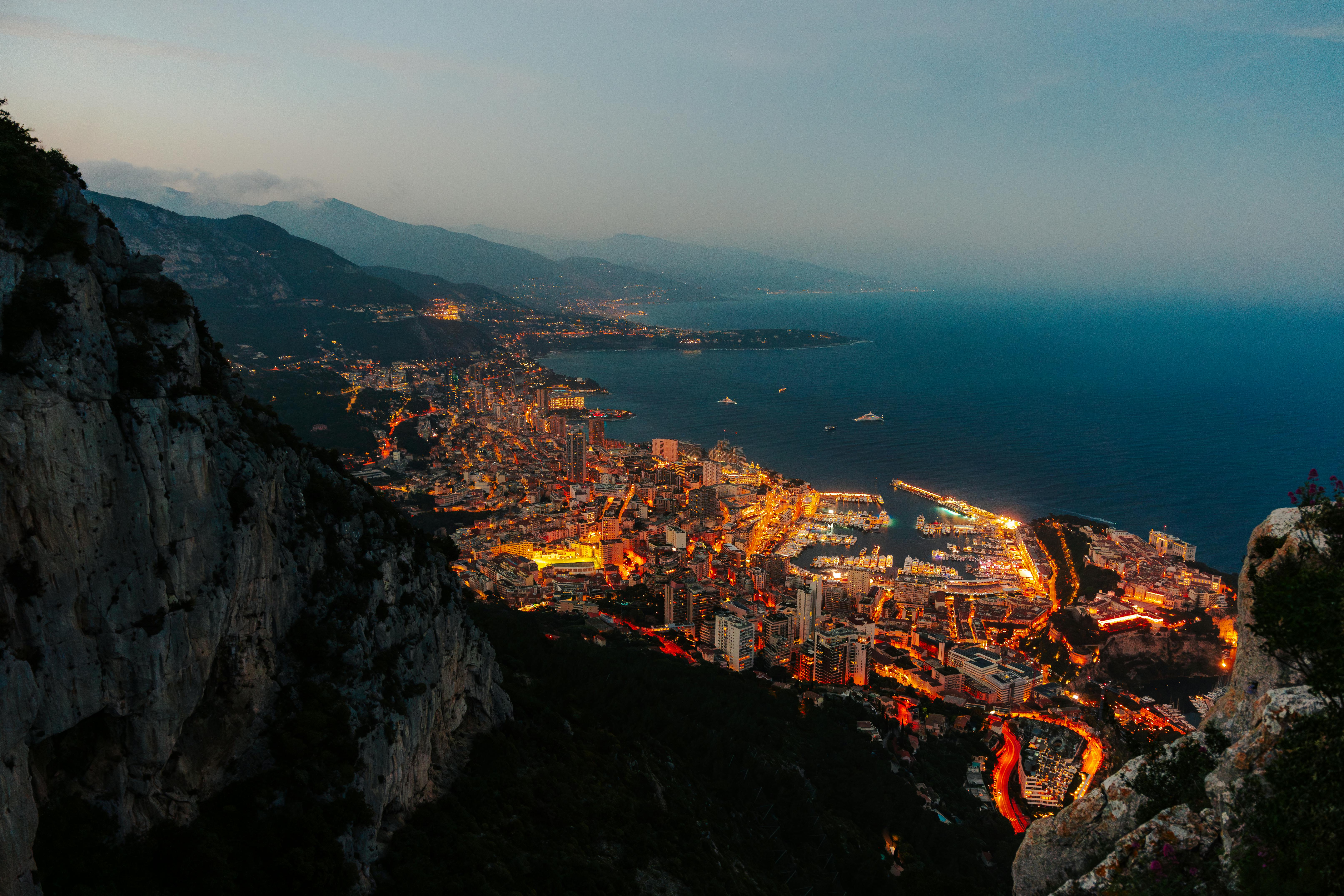 An aerial view of Monaco at dusk, with twinkling city lights and the harbour glowing against the deep blue of the Mediterranean