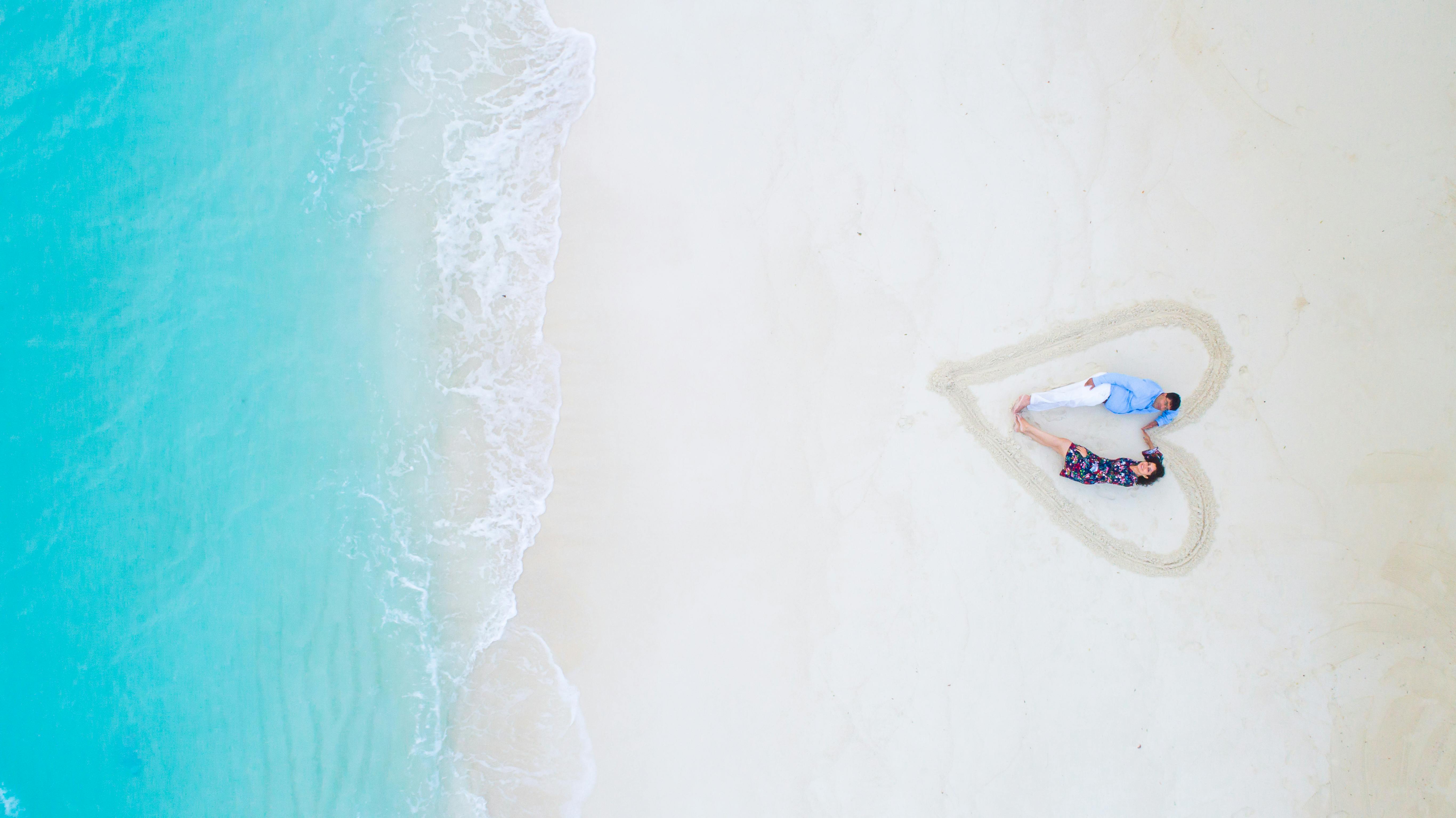 A man and woman lying on a white sand beach with a heart drawn around them