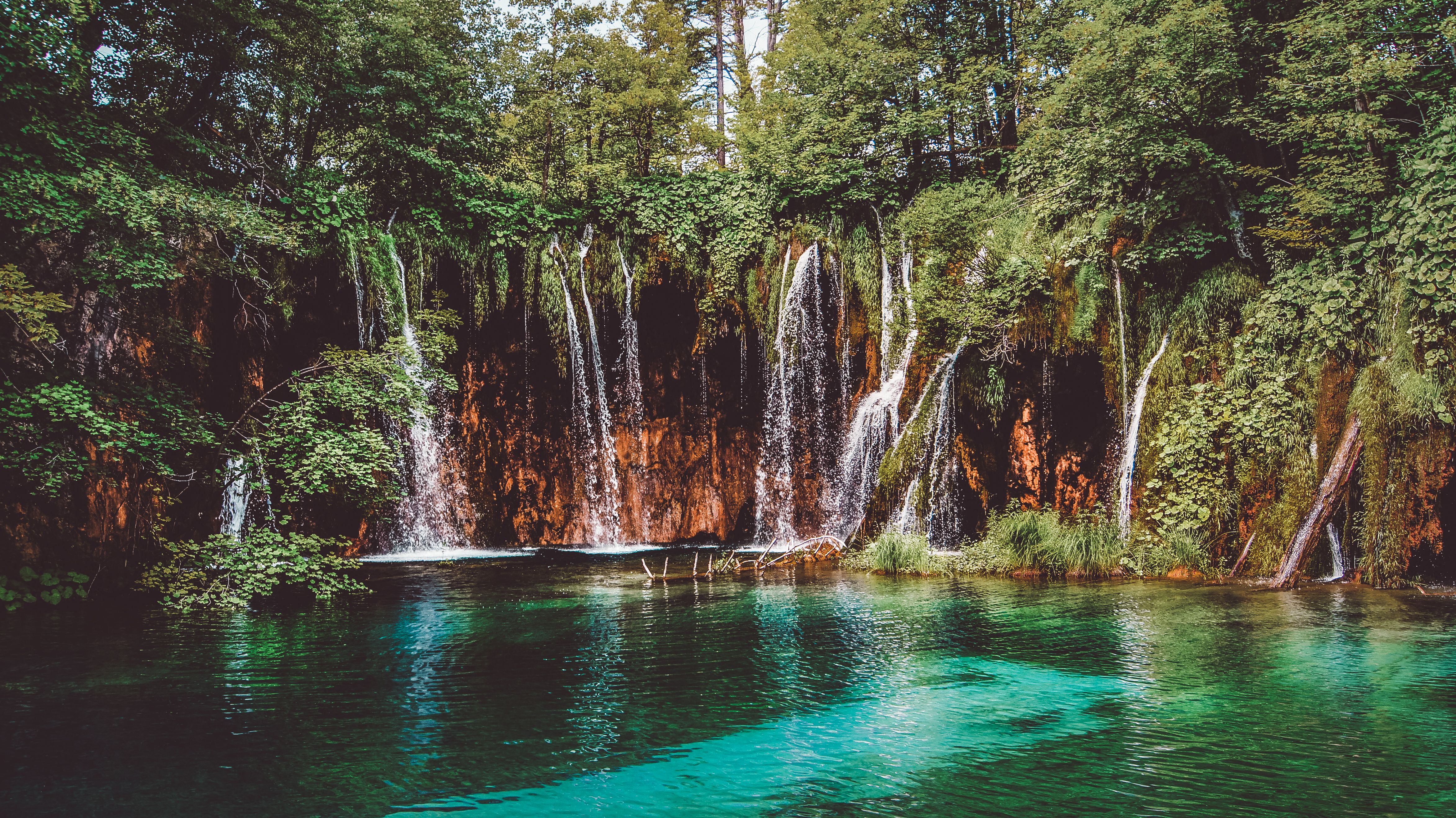 Multiple waterfalls cascade over layered limestone cliffs into turquoise pools surrounded by dense green forest